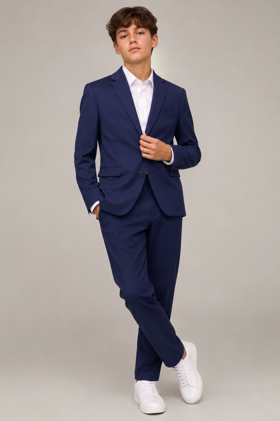 A young man wearing a navy blue two-piece suit with a white dress shirt and white sneakers, standing against a neutral gray studio background.
