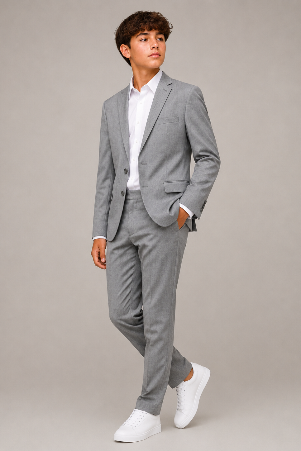 A young man modeling a light gray two-piece suit with a white dress shirt and white sneakers, standing against a solid light gray studio background.